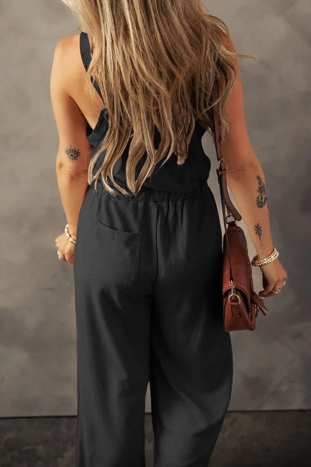 Black textured jumpsuit with knotted straps, model back view with shoulder bag