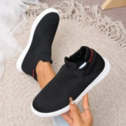 Black knit slip-on shoe held beside a matching shoe worn on a foot