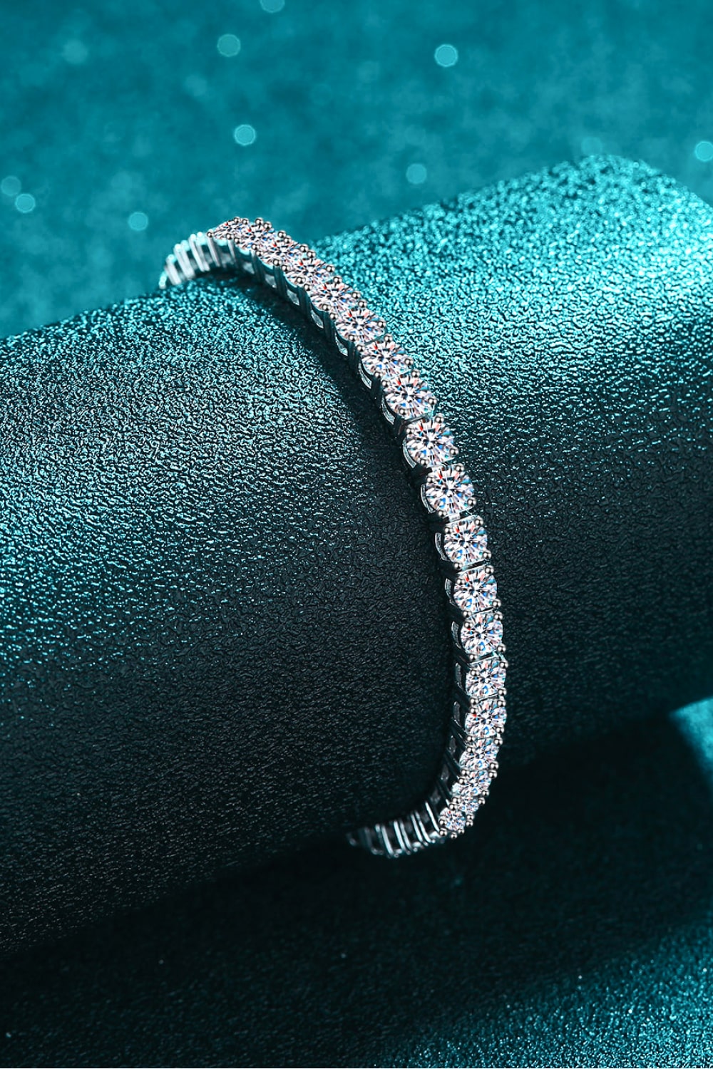 Slim silver moissanite tennis bracelet displayed on a teal velvet stand.
