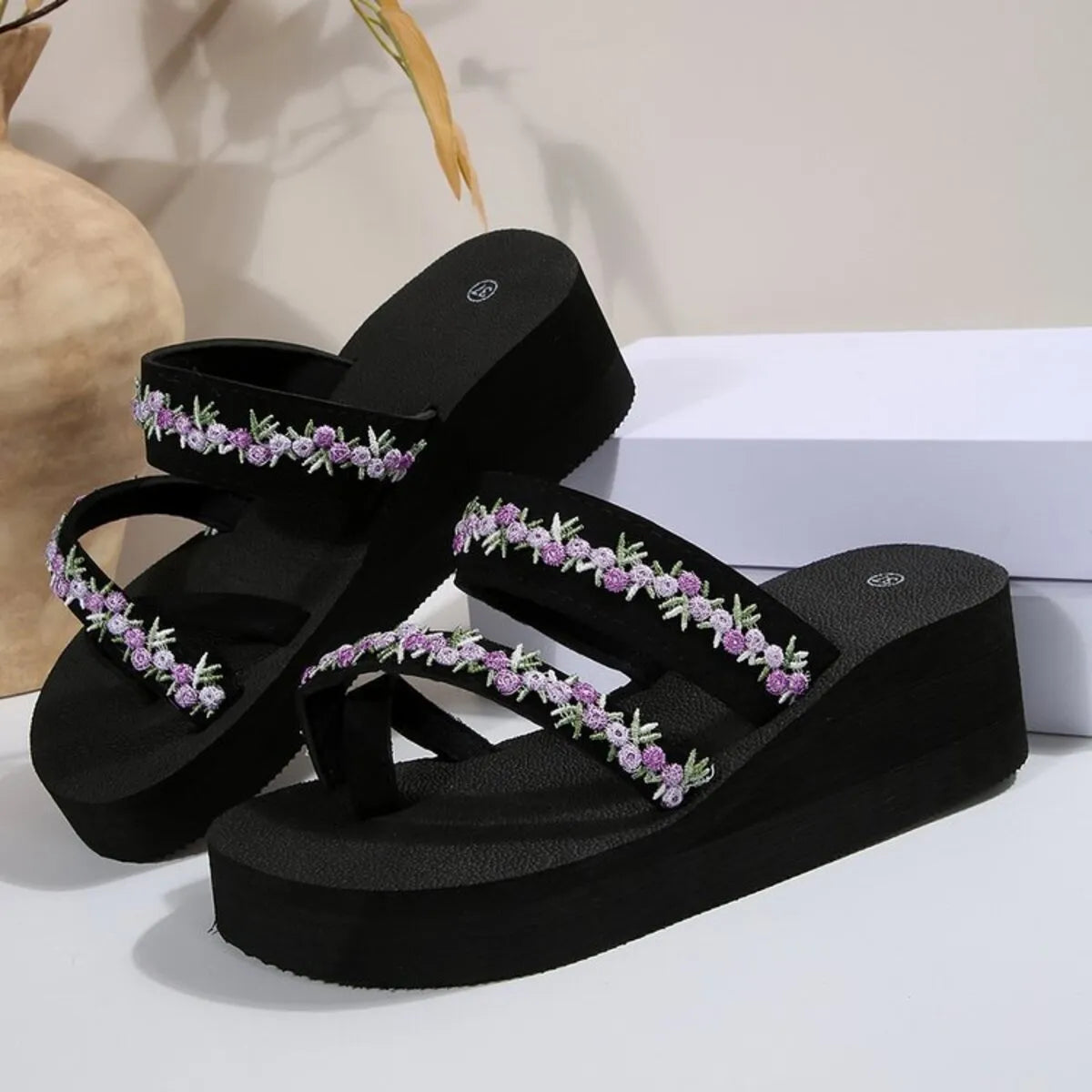 Pair of black platform sandals with purple floral-trim straps displayed on white blocks