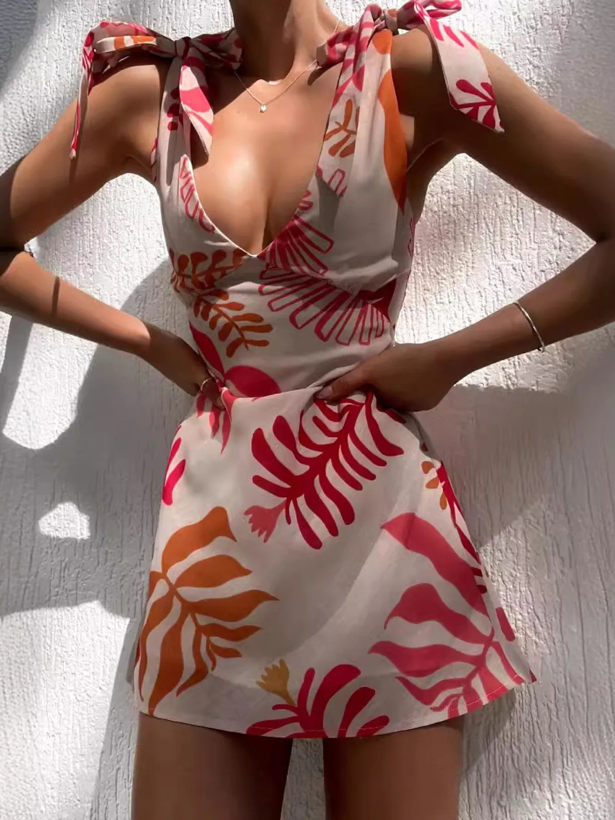 Model adjusts the tie straps on a coral-and-orange botanical print mini dress.