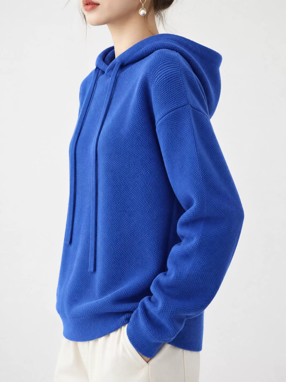 Angled view of the royal blue sweater with one hand in a white trouser pocket.