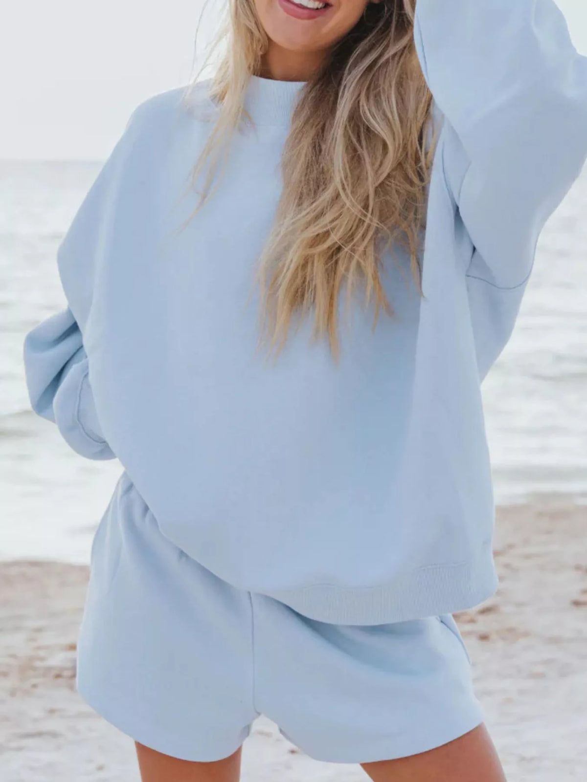 Model in a light blue sweatshirt and matching shorts at the beach, with one arm raised.