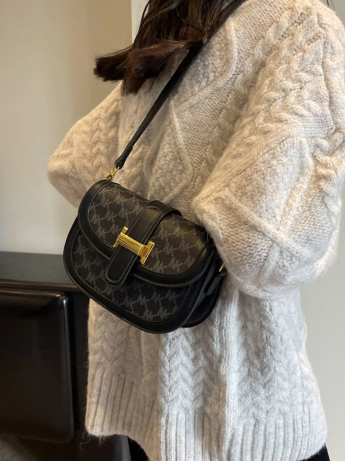 Black patterned flap crossbody bag with gold-coloured bar clasp, worn with a cream cable-knit jumper