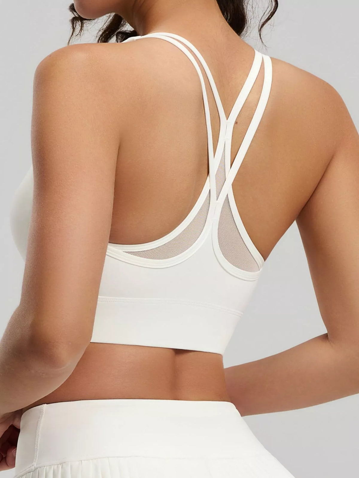 Rear angled view of white active bra with crossed double straps above a white pleated skirt