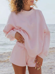 Model wearing a pale pink oversized sweatshirt and matching shorts at the beach, one hand at her waist.