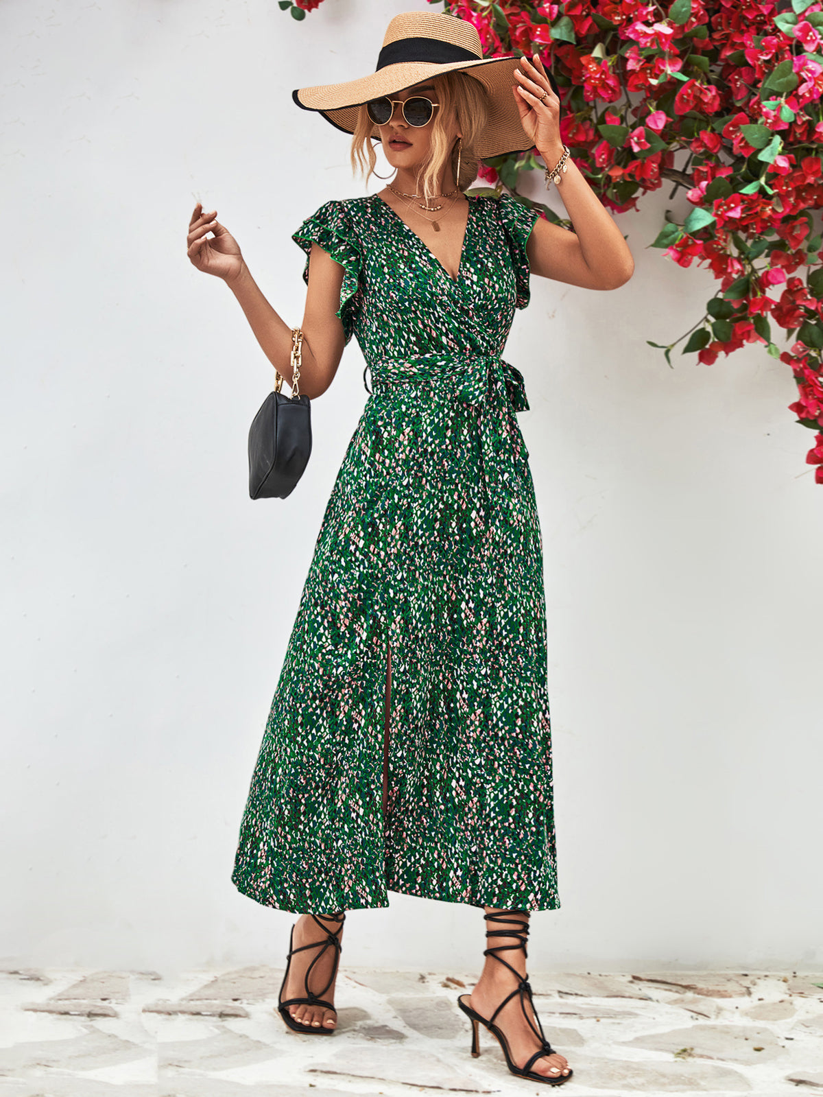 Model in green printed dress posing with a small handbag beside flowering shrubs.