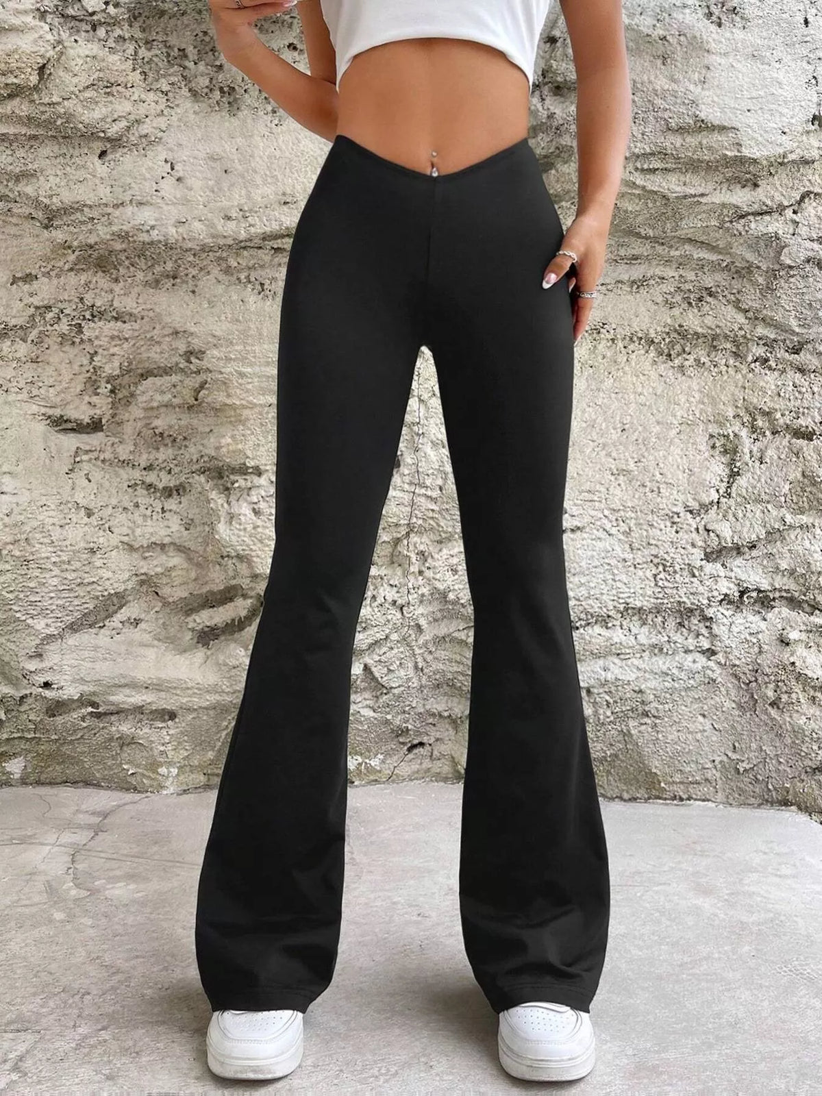 Black flared pants with a V-shaped waistband, front view with one hand at the hip against a stone wall.