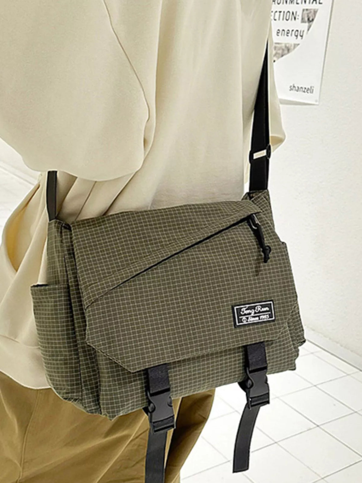 Olive fine-check messenger bag with side pockets and two buckle straps, worn with tan trousers