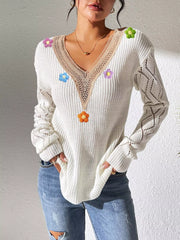 White V-neck sweater with beige trim, colourful flower appliqués and openwork sleeves, hands-in-pockets pose.