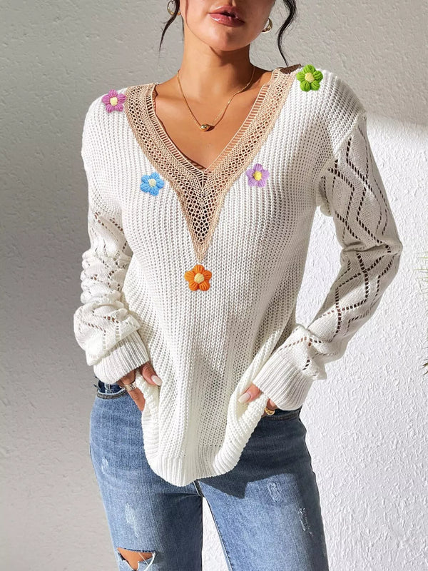 White V-neck sweater with beige trim, colourful flower appliqués and openwork sleeves, hands-in-pockets pose.