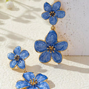 Blue flower dangle earrings on a textured pale surface with leaf-shaped shadows.