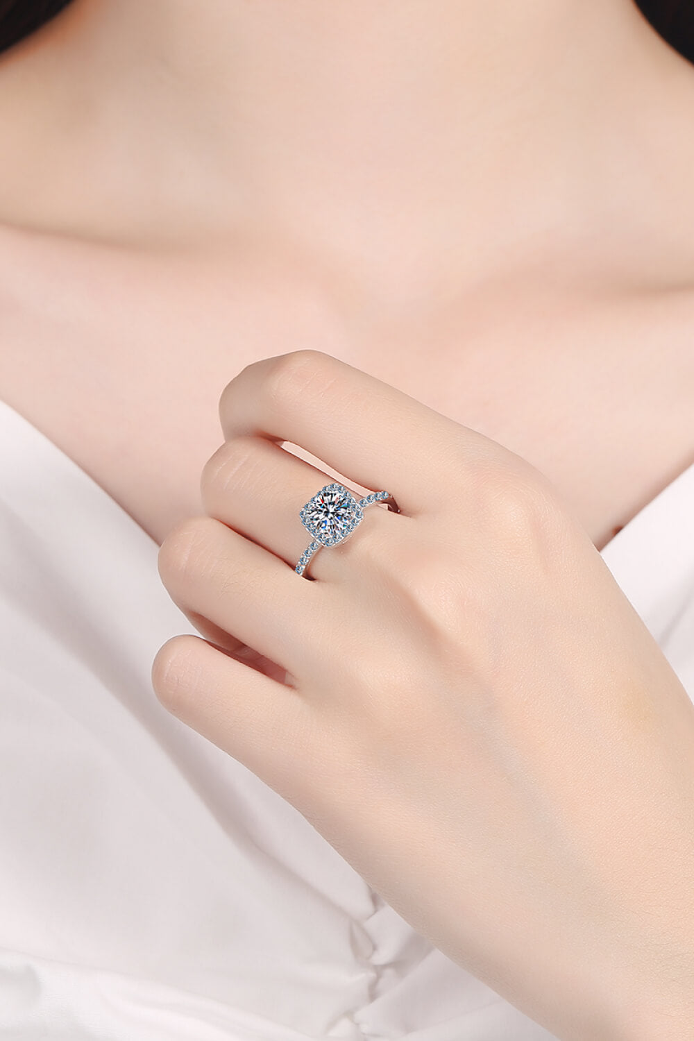 Square halo moissanite ring worn on a hand against white fabric.