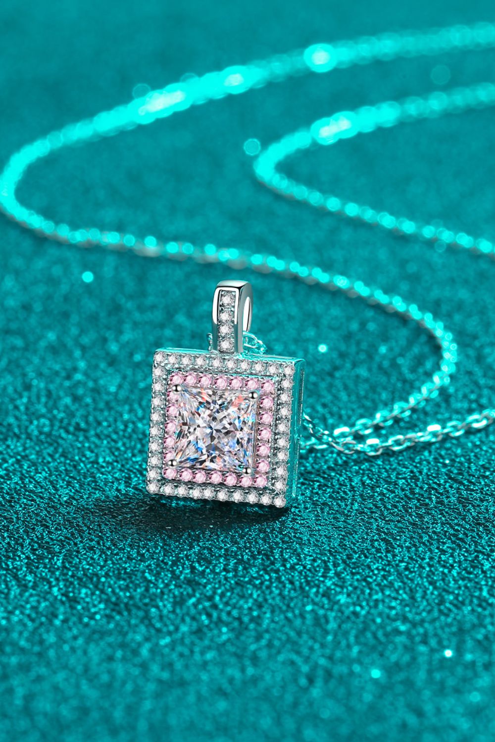 Angled view of a square pink-bordered pendant and silver-tone chain on teal