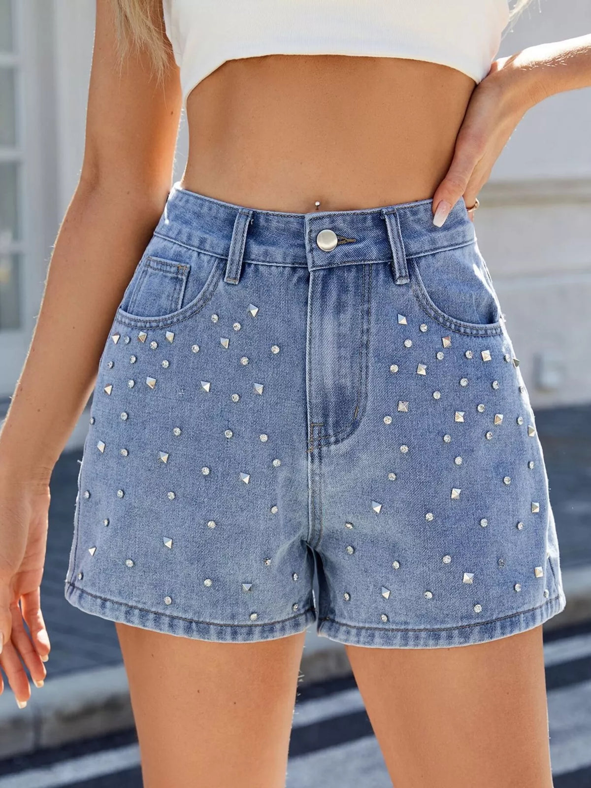 Blue high-waisted denim shorts with scattered silver-coloured studs on the front, worn with a white crop top
