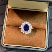 Oval royal-blue sapphire ring with a halo of clear stones, displayed in a tan jewelry box at a closer angle