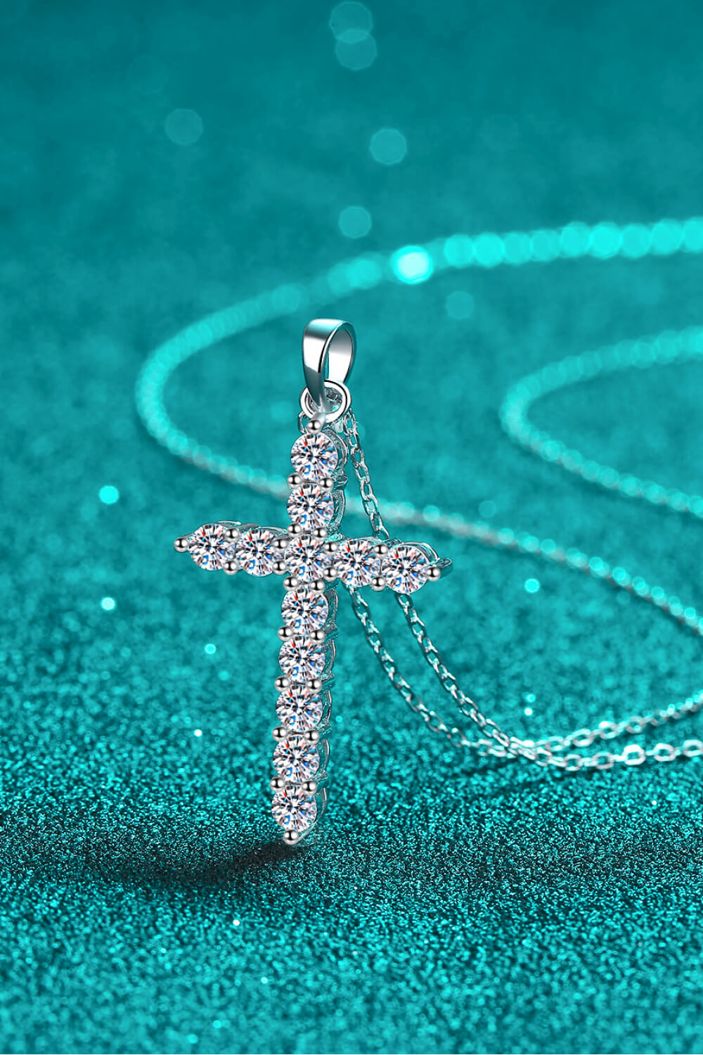 Close-up of a clear-stone cross pendant and chain resting on teal