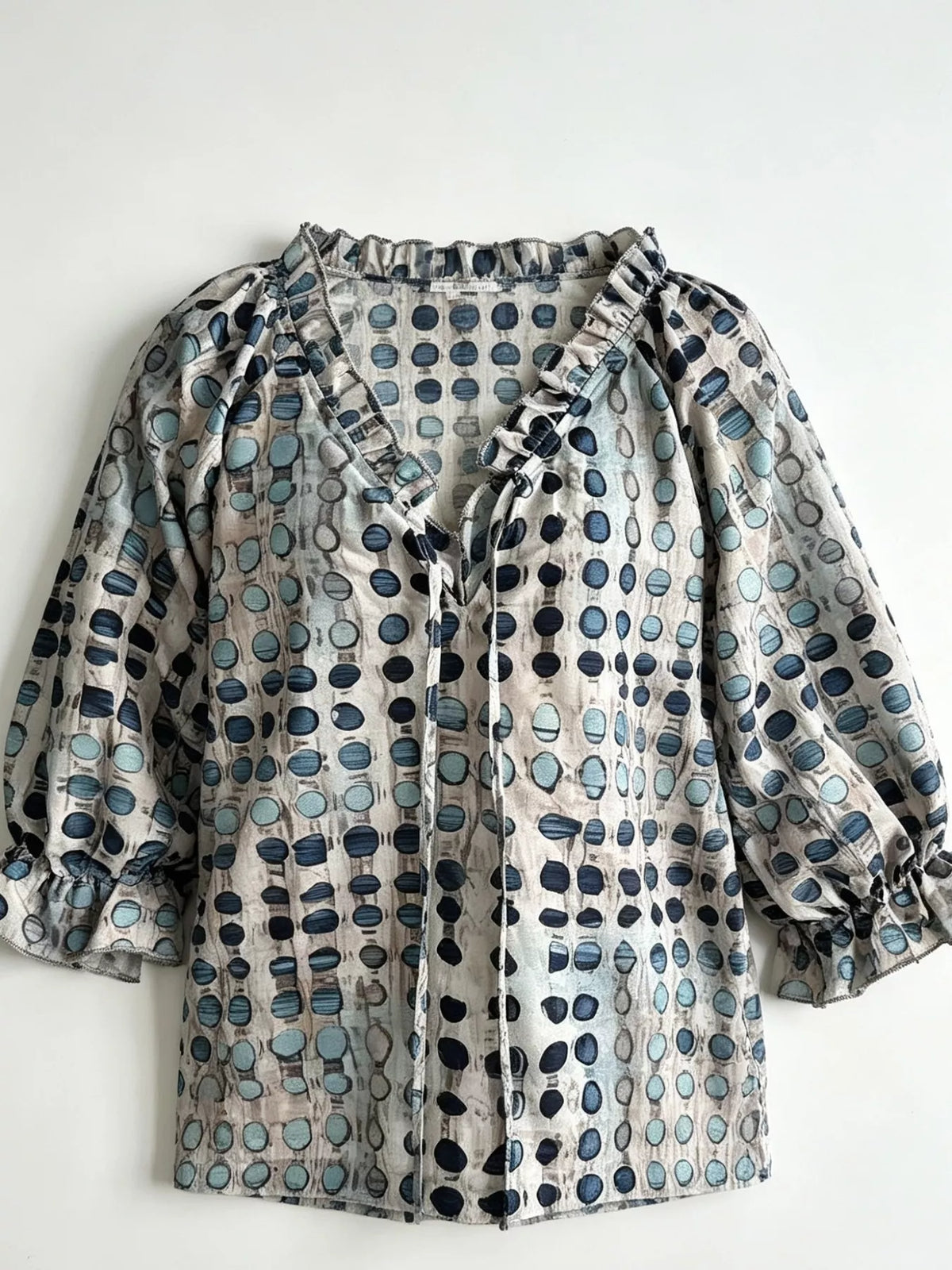 Beige blouse with blue geometric motifs laid flat, showing tie front and ruffled cuffs.