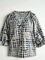 Beige blouse with blue geometric motifs laid flat, showing tie front and ruffled cuffs.