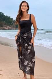 Model wearing a black square-neck swimsuit with a dark floral sarong on the beach
