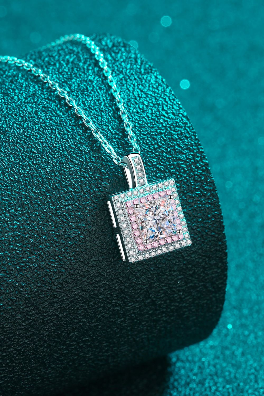 Square pink-bordered pendant necklace draped over a teal cushion
