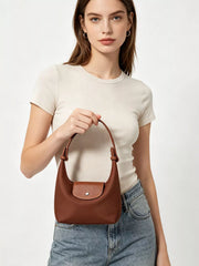 Model holding the short handle of the brown bag in front, showing its textured flap and metal snap.
