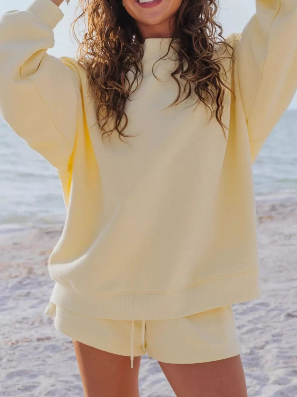 Model in a pale yellow sweatshirt and matching shorts with both arms lifted.