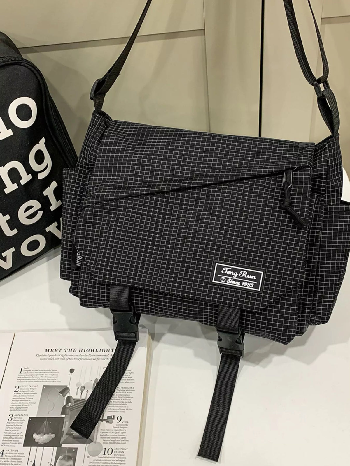 Black checked messenger bag with front zip pocket, side pockets and hanging buckle straps, displayed against white