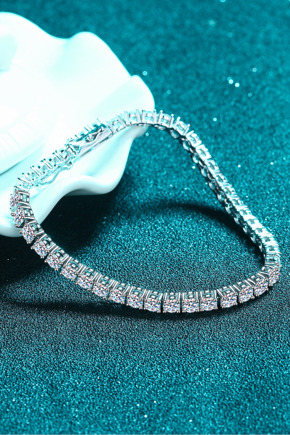 Curved silver tennis bracelet displayed on teal fabric beside a white flower.