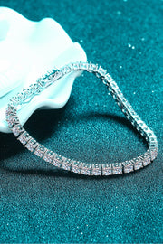 Curved silver tennis bracelet displayed on teal fabric beside a white flower.