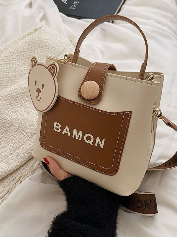Ivory bucket bag with brown trim, bear charm and shoulder strap displayed at an angle