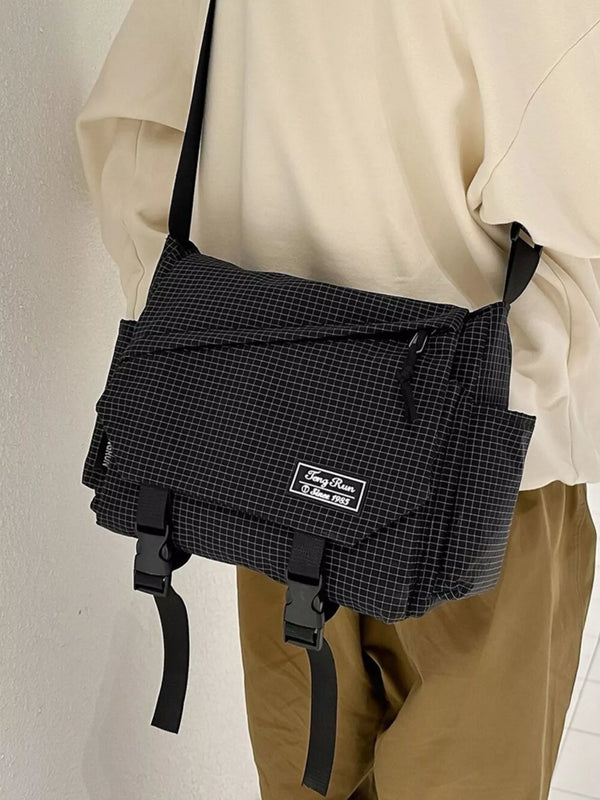 Black fine-check crossbody messenger bag with front flap, diagonal zip pocket and two buckle straps, worn at the hip