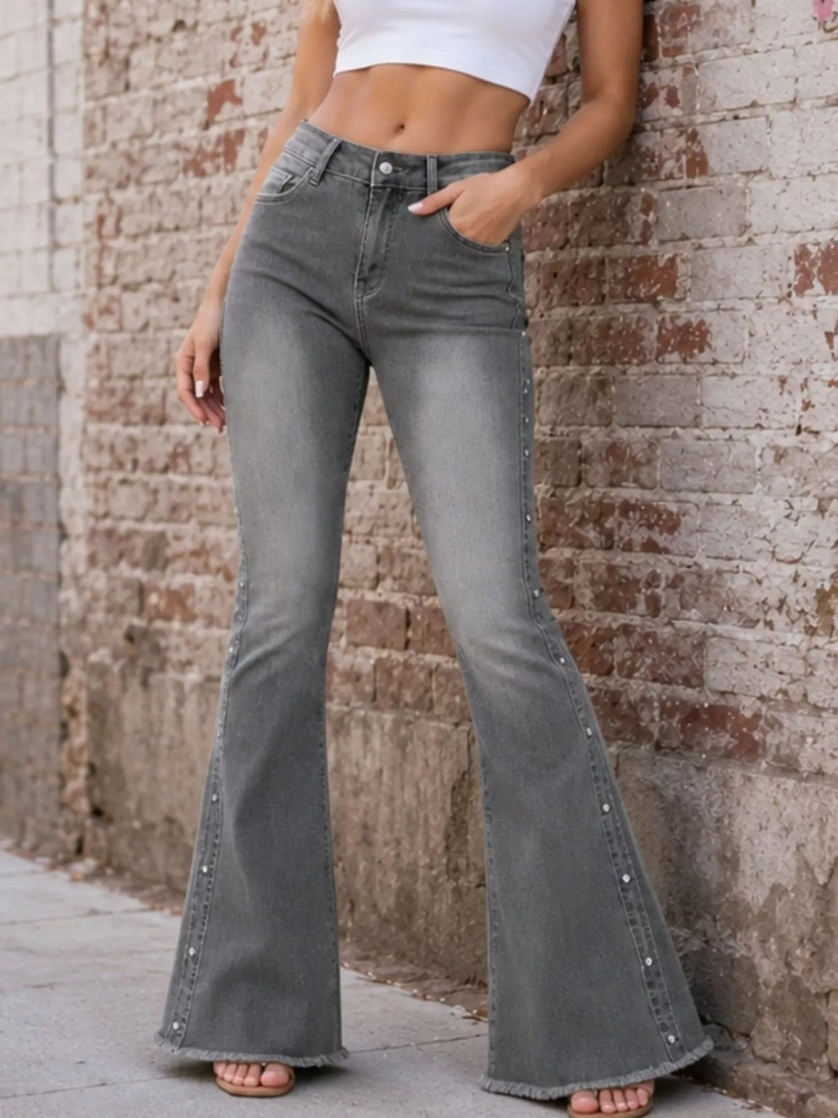 Grey high-waisted flare jeans with studded outer seams styled with a white crop top by a brick wall