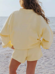 Back view of a pale yellow sweatshirt and shorts set on a model with hands at her hips.