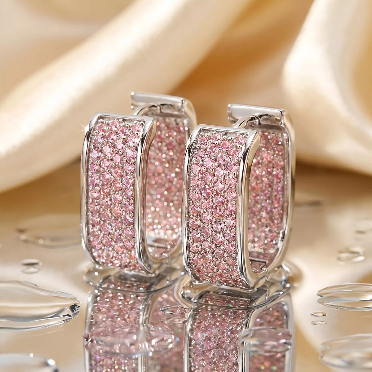 Silver-tone square hoop earrings with pink stones on reflective surface