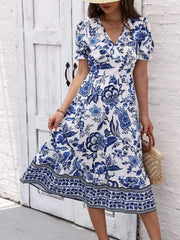 Blue floral midi dress with short puff sleeves and a border-print hem, shown holding one side of the skirt.