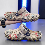 Beige graffiti-print clogs with wide instep straps, one held above a matching shoe