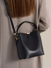Black PU leather-look bucket bag worn crossbody over a taupe coat, with its short handle and gold-tone hardware.