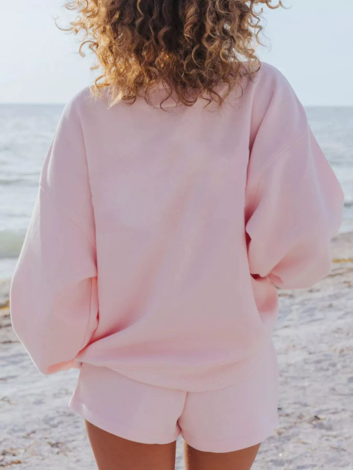 Back view of a pale pink crewneck sweatshirt and matching shorts on a model at the beach.