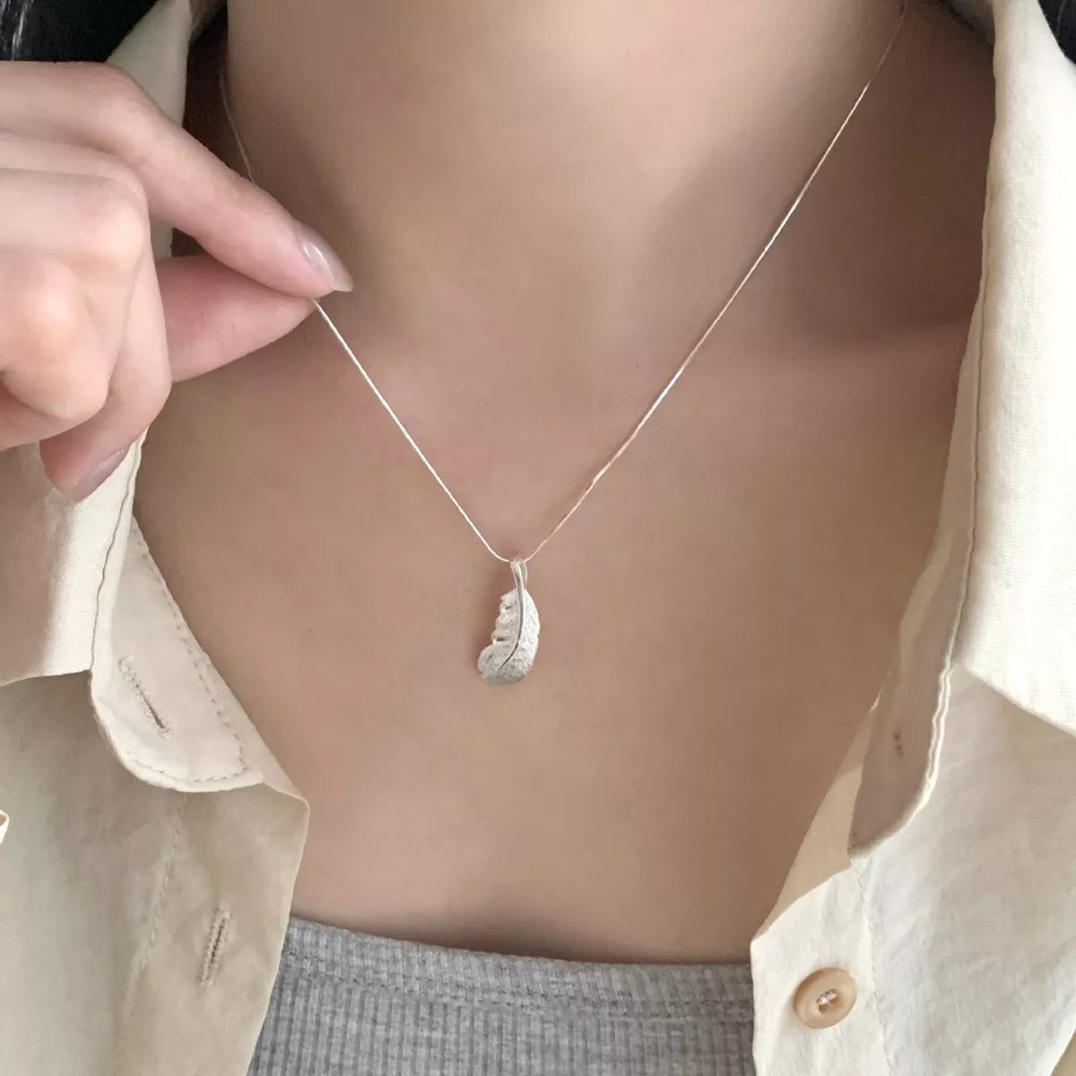 Model holding a silver feather pendant necklace against a cream shirt