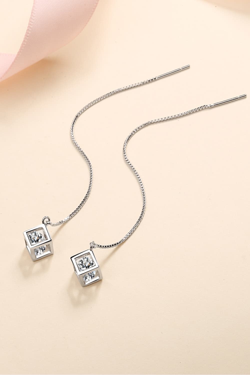 Pair of square-drop threader earrings laid across a peach-coloured background