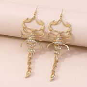 Pair of gold-tone scorpion dangle earrings with clear accents on a wrist