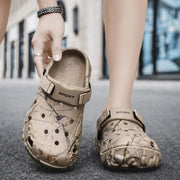 Khaki branch-print clogs with one worn and one held to show the footbed