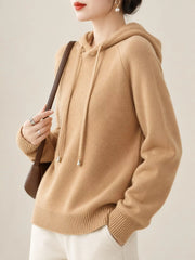 Camel hooded sweater shown at an angle with one hand in a white trouser pocket.