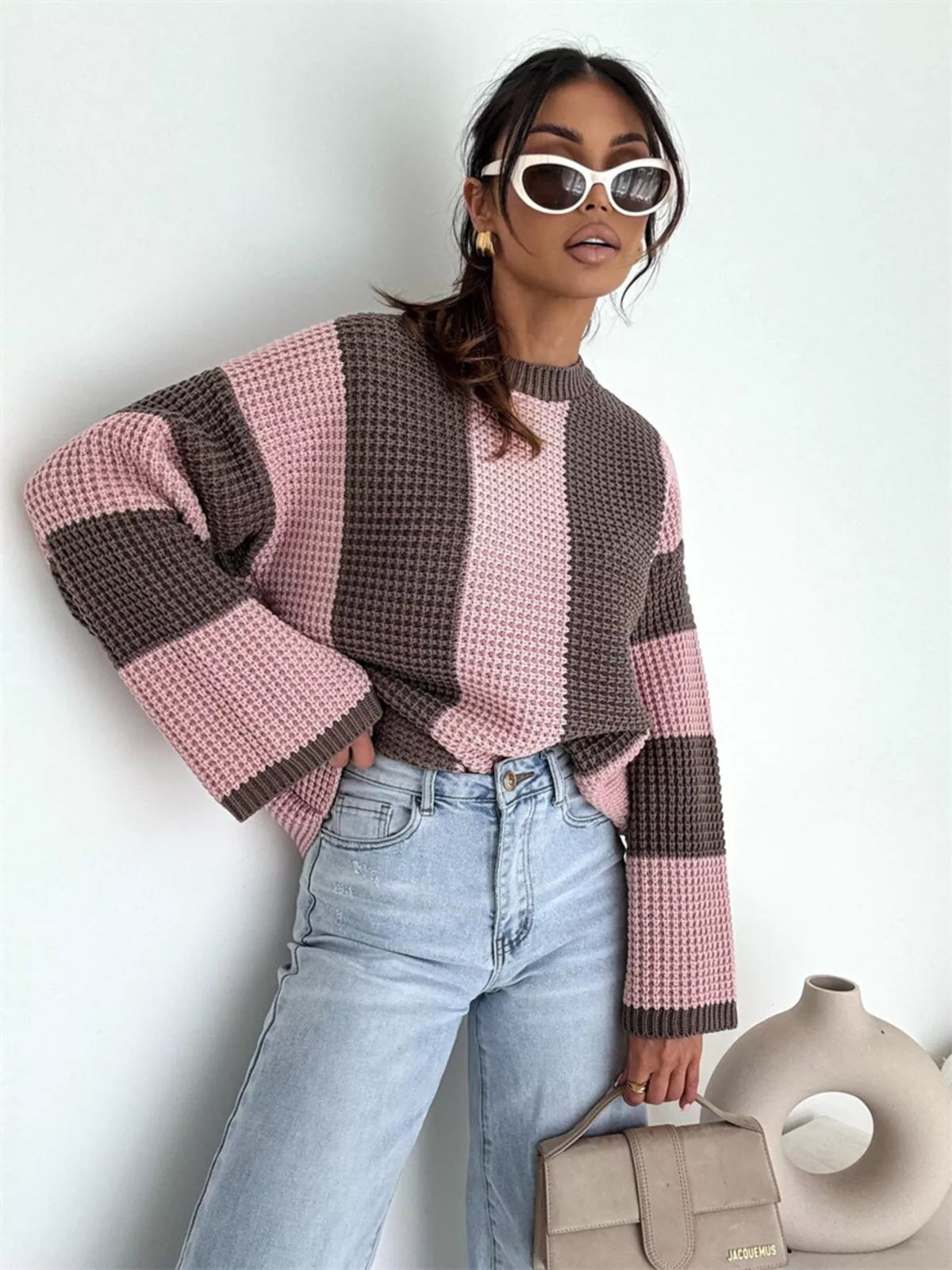 Model wearing the pink and brown sweater seated with one knee raised