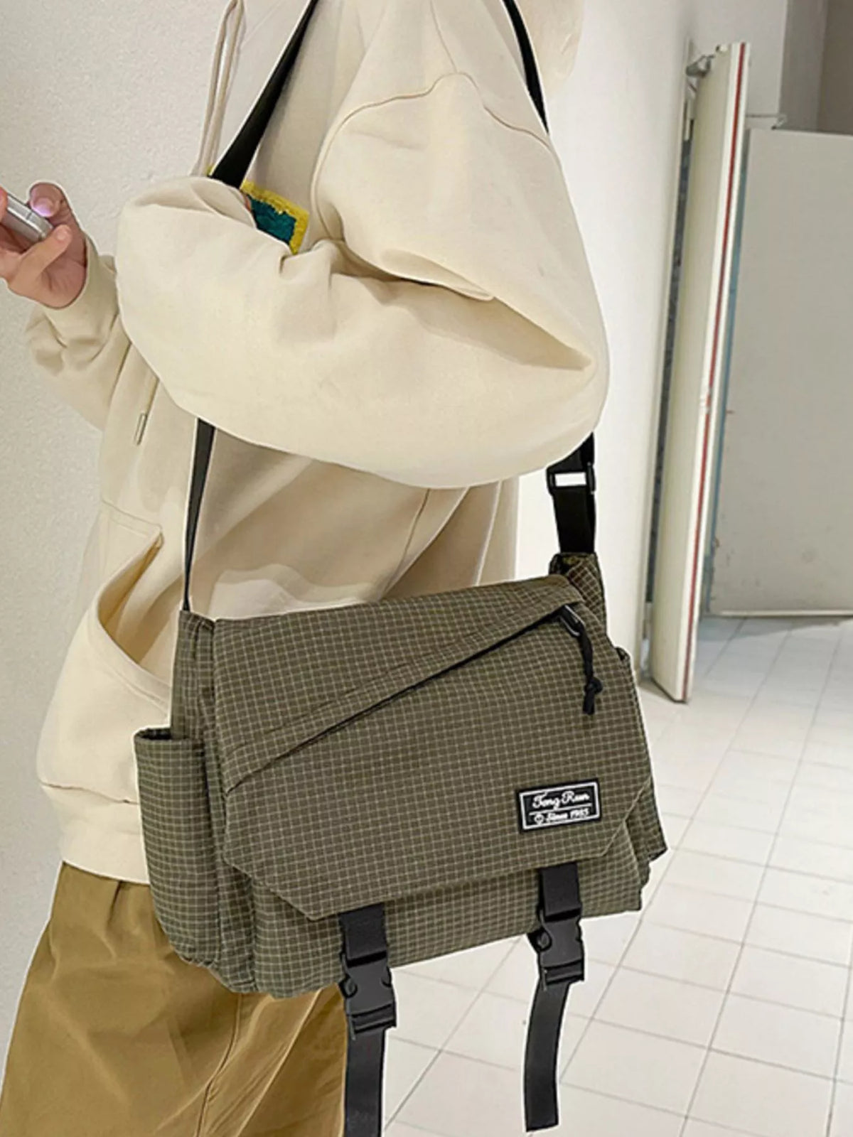 Side view of olive fine-check shoulder bag with flap, zip pocket and hanging buckle straps