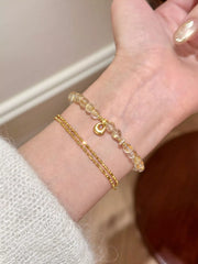 Gold charm bracelet and fine chain bracelet worn together on a wrist.