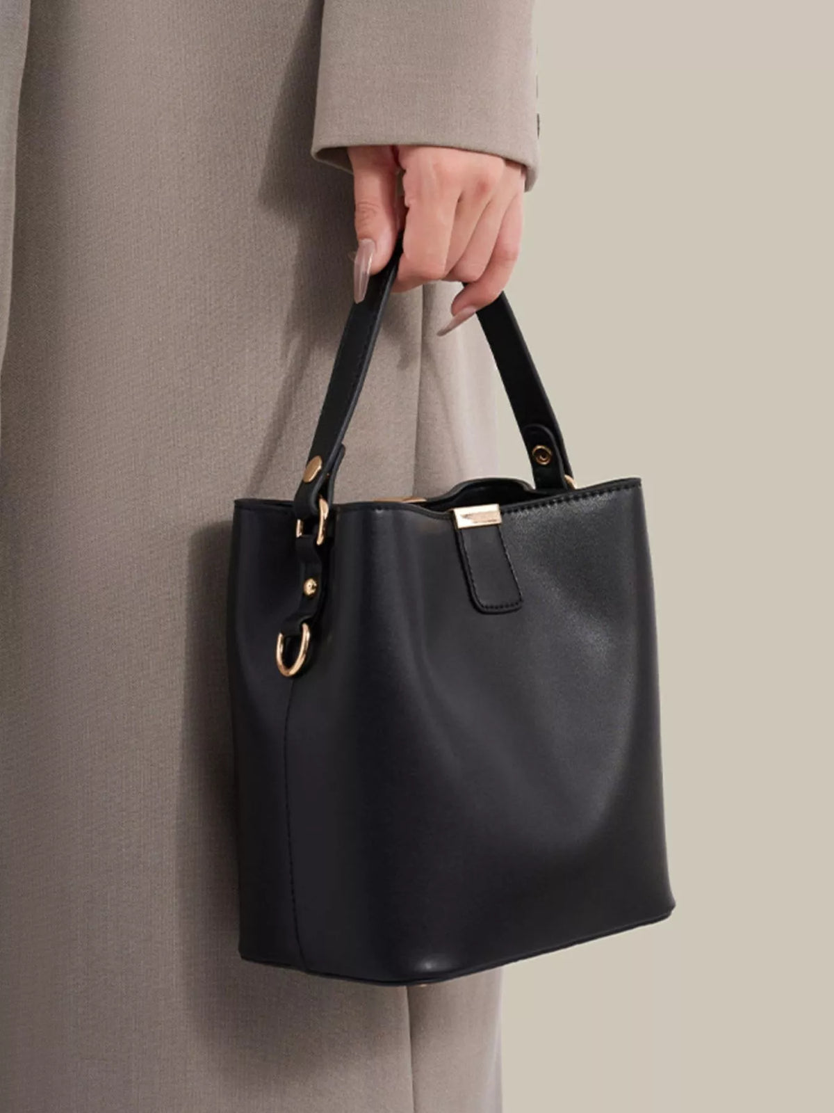 Black bucket bag held by its short handle, with the long detachable strap and gold-tone side clips visible.