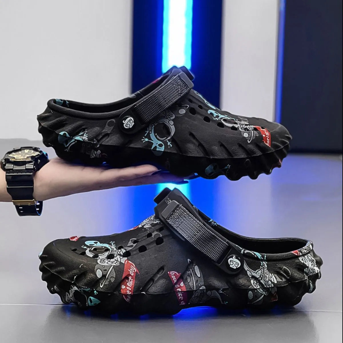 Black graffiti-print clogs with instep straps and sculpted soles, one held above a matching shoe