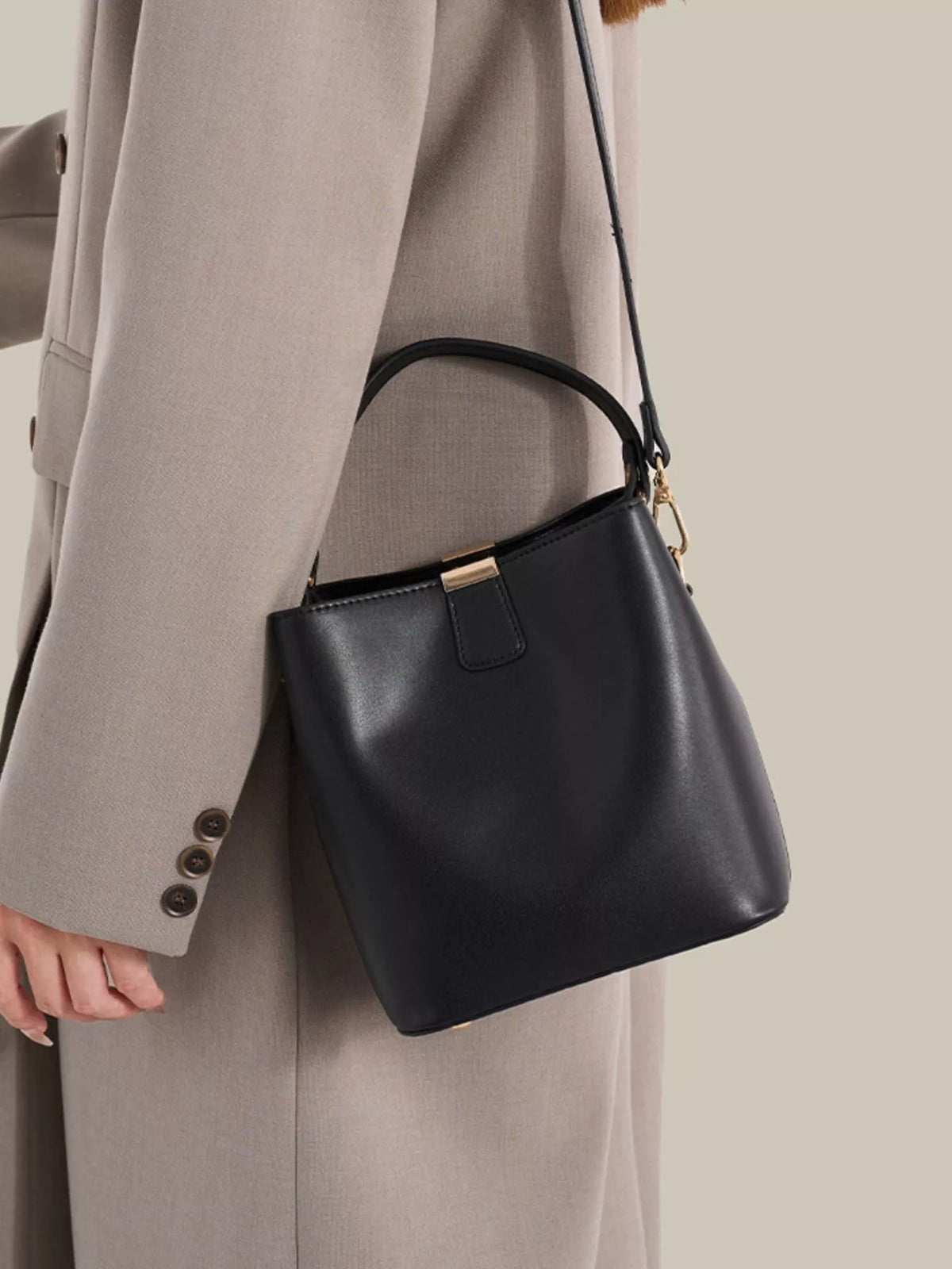 Black PU leather-look bucket crossbody bag worn against a taupe coat, showing the gold-tone clasp and short handle.